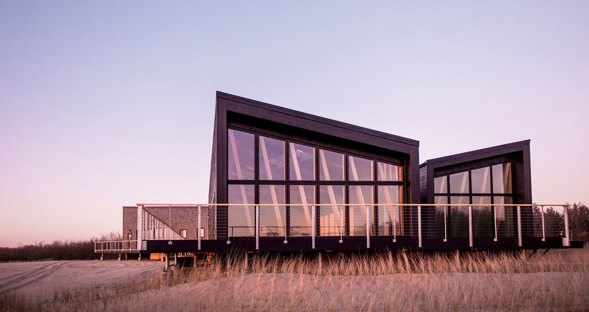 The image shows a modern building with large windows and a slanted roof, set against a sunset sky, surrounded by tall grass.