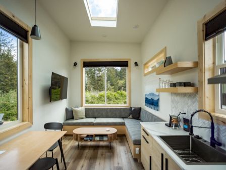 A modern tiny house interior with a kitchenette, cozy seating, and large windows showcasing a lush outdoor view.