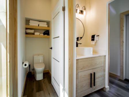 A modern bathroom with a toilet, sink with mirror, wall lights, and open shelves holding towels above the toilet.