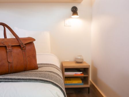 A leather bag on a bed, next to a small nightstand with books and a lamp, in a cozy room setting.