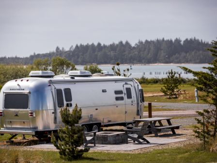 A silver Airstream trailer parked at a campsite with picnic tables, trees, and a scenic lake and forest in the background.