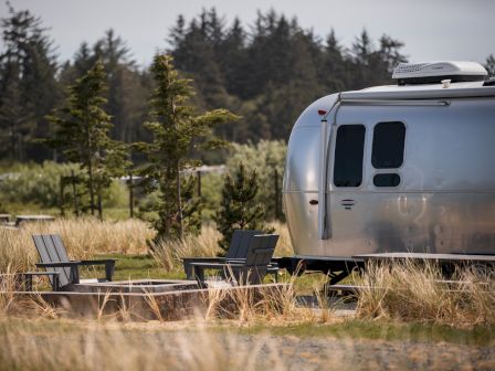 Airstream trailer with two Adirondack chairs beside a fire pit, set in a grassy area with trees in the background.