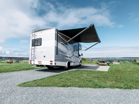 An RV parked on gravel with an awning extended, set in a grassy area near a body of water under a partly cloudy sky.