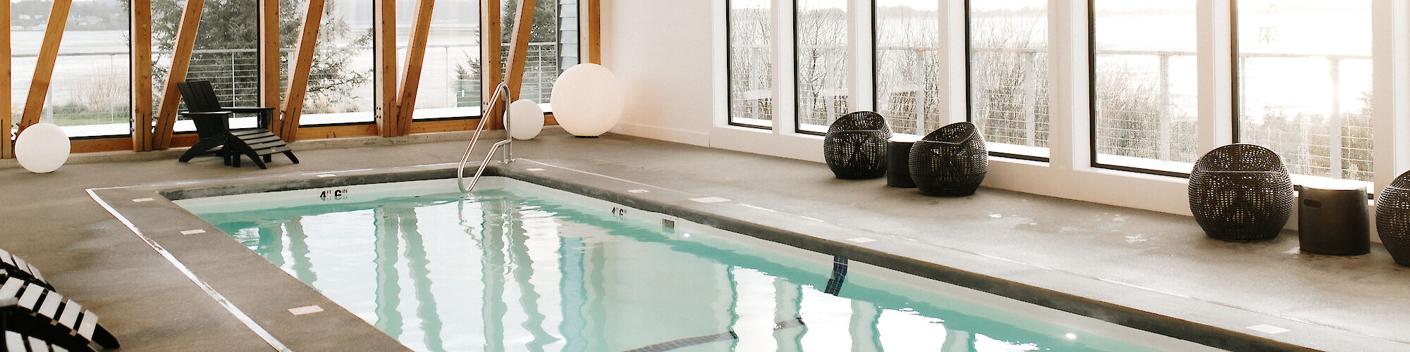 A modern indoor pool with wooden beams, lounge chairs, large windows, and spherical lights, offering a serene and stylish atmosphere.