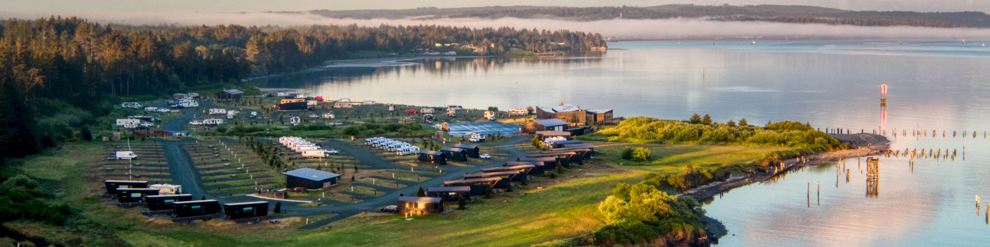 A coastal landscape with a grassy peninsula, buildings, RVs, and calm water reflecting the sky and clouds at sunset.