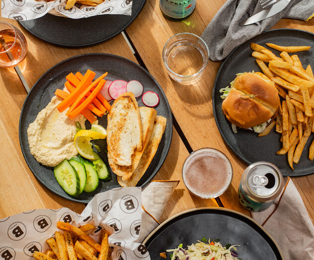 The image shows plates of food on a wooden table, including fries, burgers, wraps, veggies, and drinks.