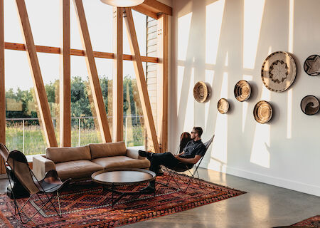 A modern living room with large windows, unique wooden beams, wall decor, and a man sitting on a chair next to a sofa and round coffee table.