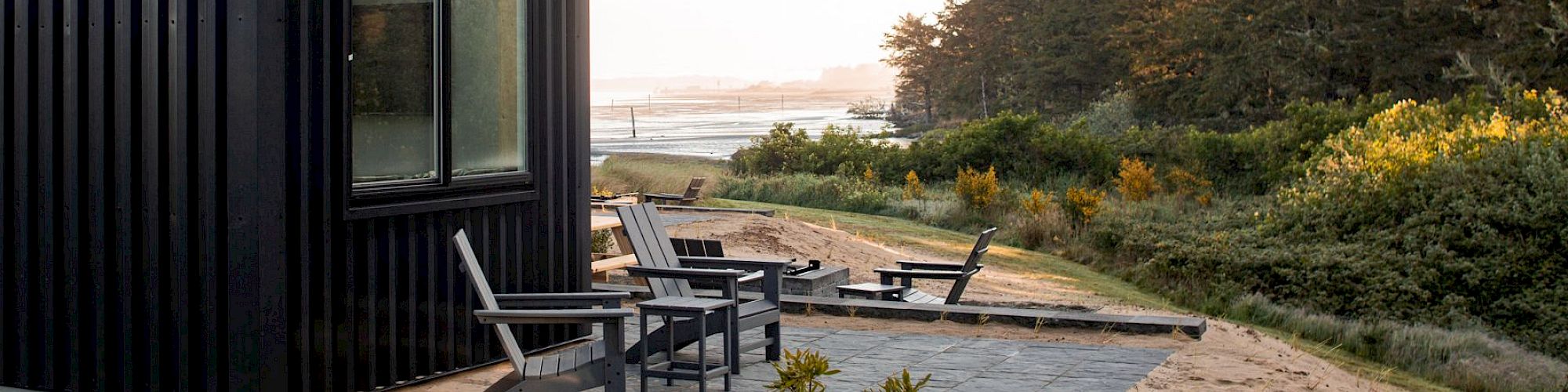 A modern black cabin with a patio and outdoor chairs, set near a forest with a view of the water in the background.