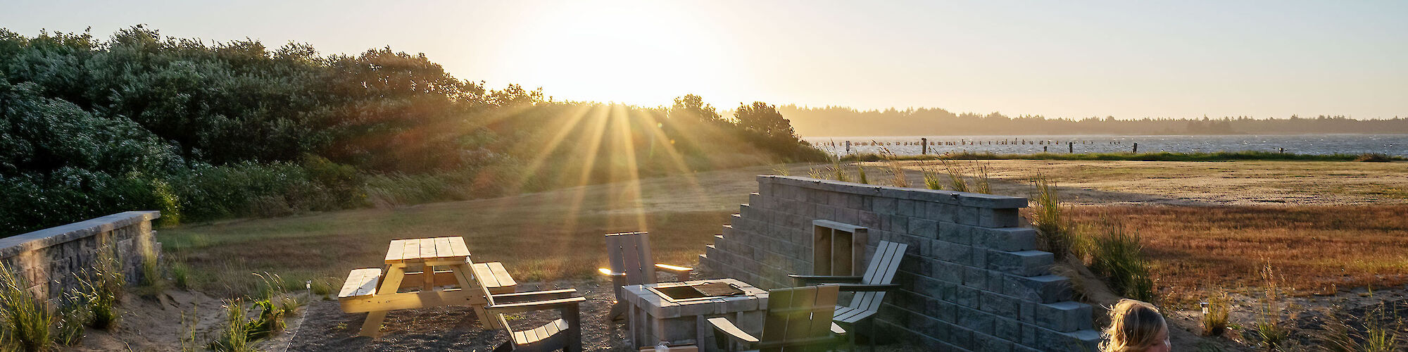 A child plays near a modern outdoor seating area at sunset, surrounded by nature and coastal views.