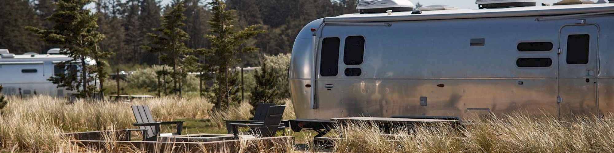 Airstream trailers are parked in a grassy area with trees and outdoor seating visible under a clear sky.