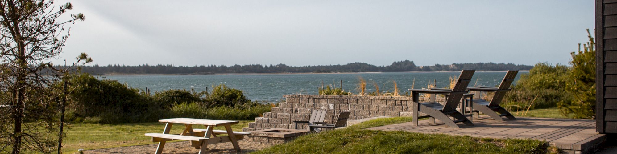 A serene coastal scene with a picnic table, wooden chairs, and lush grass overlooking the ocean under a clear blue sky.