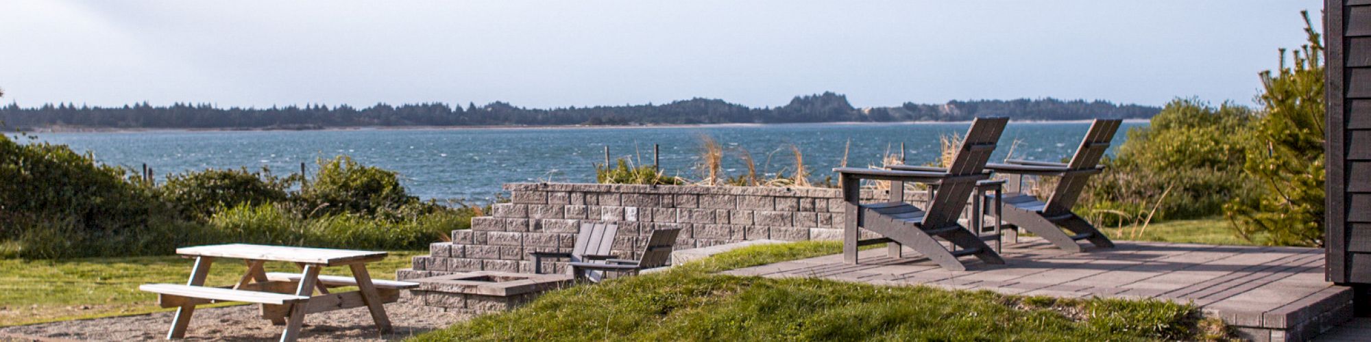 A scenic outdoor area with a picnic table, wooden chairs, and a view of the water and distant hills under a clear blue sky.