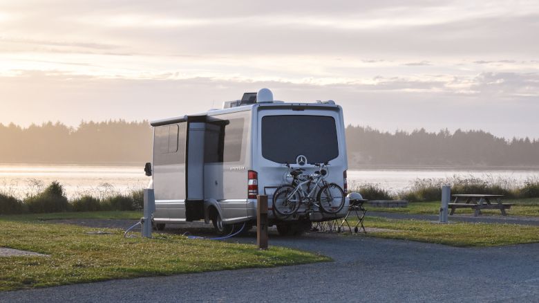 A camper van parked by a lakeside at sunset with bicycles attached to the rear. A picnic table and grassy area are visible nearby.