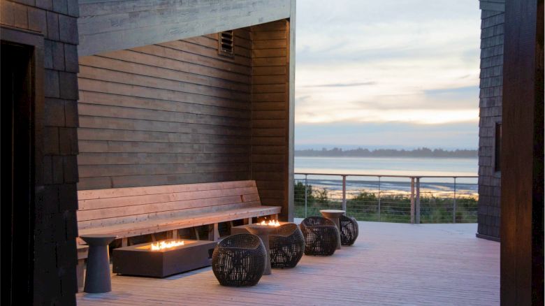 A modern outdoor seating area with wooden benches, decorative lanterns, and a view of the water in the background at dusk.