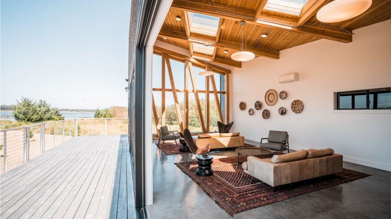 A modern living room with wooden ceiling, skylights, and outdoor view. Features cozy sofas, circular wall decor, and a spacious deck outside.