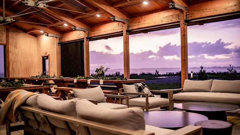 A cozy outdoor lounge with comfortable sofas and a wooden ceiling, overlooking a scenic sunset view through large open windows.