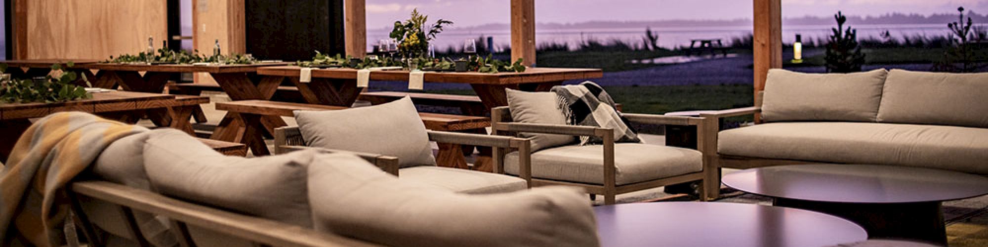 A cozy open-air room with cushioned seating, wooden beams, and a view of a purple-hued sunset sky.