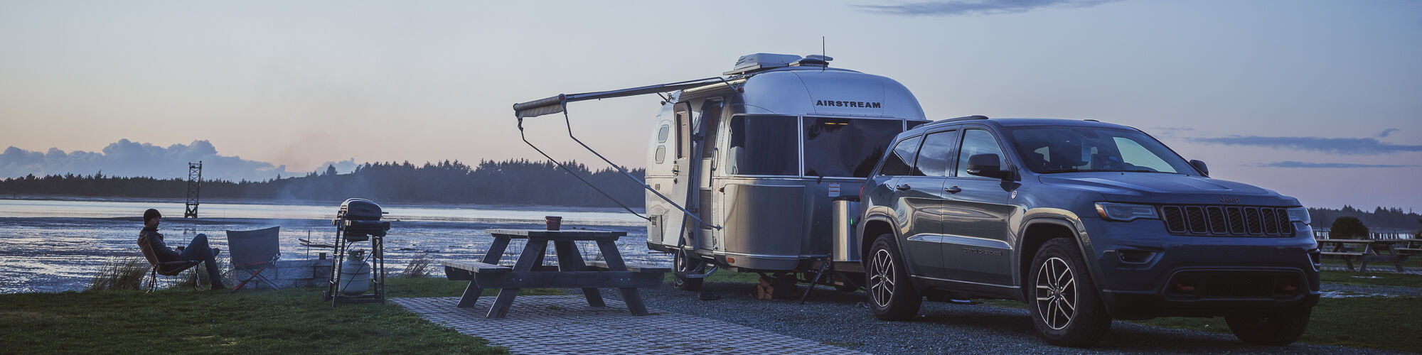 A parked SUV and travel trailer by a lake during sunset, with two people sitting near a picnic table, enjoying the view.