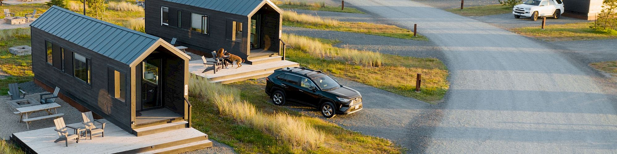 The image shows two small black cabins with decks, surrounded by grass. A black car is parked nearby on a gravel path.