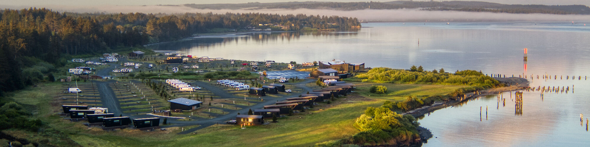 An aerial view of a campground by a calm lake, surrounded by trees and a partly cloudy sky.