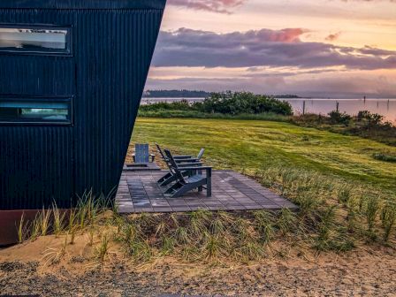 A modern building with dark siding overlooks a grassy landscape and water, beneath a colorful sunset sky.