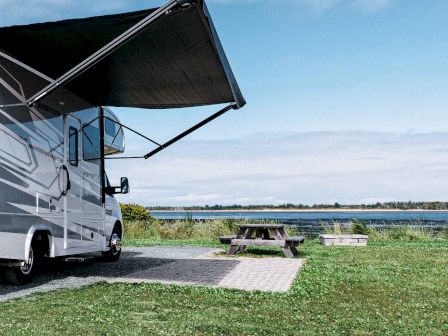 A motorhome with an extended awning is parked near a grassy area by a lake, with a picnic table and clear blue sky in the background.