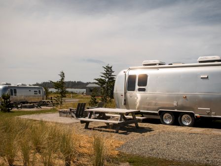 Two silver RVs parked at a campsite with picnic tables and chairs, surrounded by grass and trees under a cloudy sky.