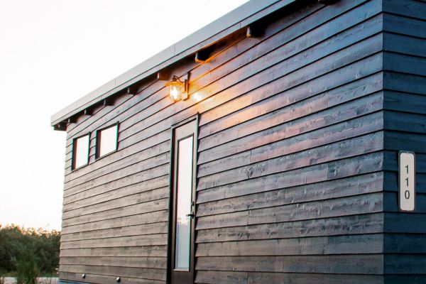 A small, modern, black wooden house with a slanted roof and a lit exterior light, located on a sandy landscape with a gravel path.