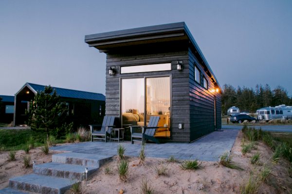 A modern tiny house with large windows, patio chairs, and steps leading up to it; nearby trees and trailers are visible in the background.