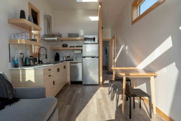 A modern kitchen and dining area with wooden cabinets, stainless steel appliances, a table with stools, and a sofa. Sunlight streams in.