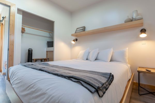 A cozy bedroom with a double bed, white linens, a striped throw, a shelf with decor, and a small side table is shown.