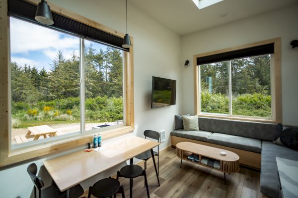 The image shows a cozy room with large windows, a dining table, chairs, a wall-mounted TV, and a comfortable seating area with outdoor views.