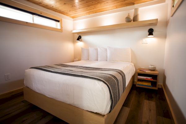 A cozy bedroom features a neatly made bed with a striped blanket, wooden accents, a small shelf, and warm lighting from wall sconces.