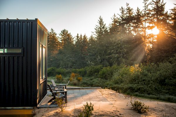 A modern black cabin with patio chairs is surrounded by lush greenery and trees, basking in a beautiful sunrise.