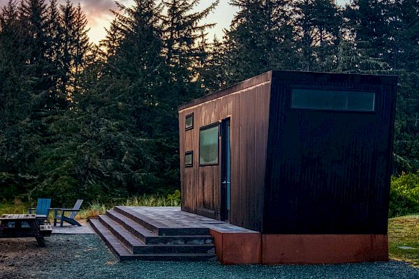 A modern cabin with steps is in a forest setting. Nearby, there's a picnic table and chairs on a deck at sunset.