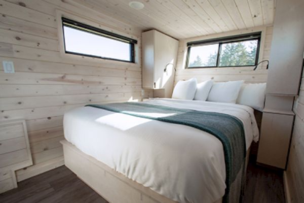 A cozy bedroom with wood paneling features a neatly made bed, two windows, and built-in lighting, creating a warm and inviting space.