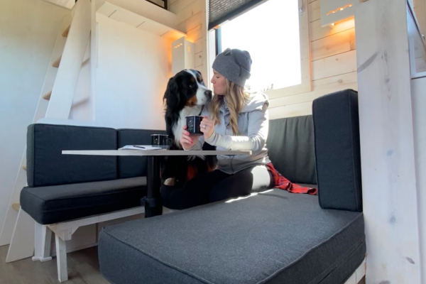 A person and a dog are sitting at a small table in a cozy, modern space with lofted beds and wooden walls. The scene is well-lit.