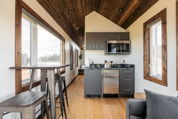 Modern tiny house interior with a small kitchen, wood accents, and windows. High table with stools and a cozy sitting area are visible.