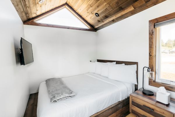 A cozy bedroom features a bed, a mounted TV, and a window, all under a wooden ceiling with a triangular skylight.