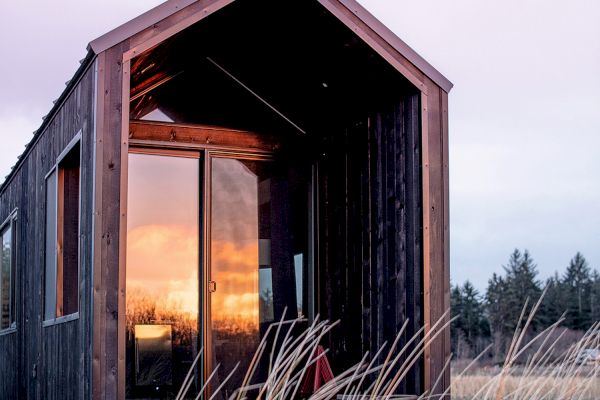 A small, modern cabin with a wooden exterior shows a sunset reflected in its glass door, surrounded by tall grasses and distant trees.