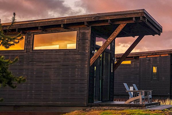 A modern wooden cabin with large windows, featuring two chairs on a small porch, set against a sunset sky.