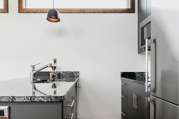 A modern kitchen with grey cabinets, marble countertops, stainless steel appliances, wooden flooring, and pendant lighting hangs from wooden beams.