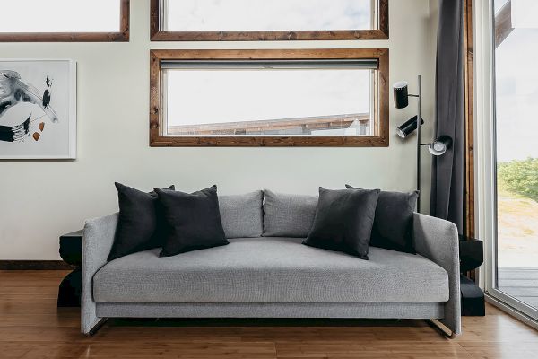 The image shows a modern living room with a gray sofa, black cushions, framed artwork, large windows, and a floor lamp against a wooden floor.