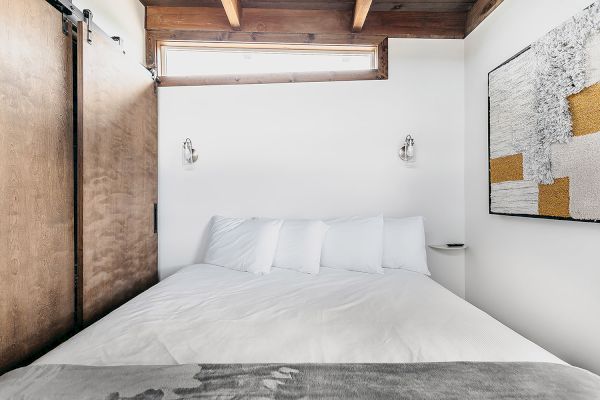 A cozy bedroom with a wooden ceiling, white walls, a bed with white linens, wall lights, and abstract artwork on the wall.