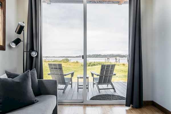 A cozy room with a sofa leads to a patio through sliding glass doors, featuring two chairs and a scenic view outside.