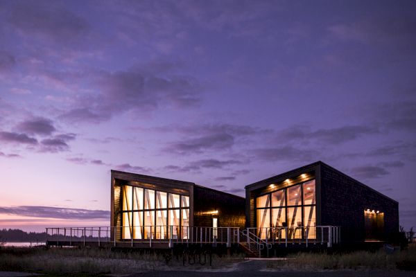 A modern building with large windows is illuminated under a purple sky at dusk, featuring a scenic outdoor view and warm interior lights.