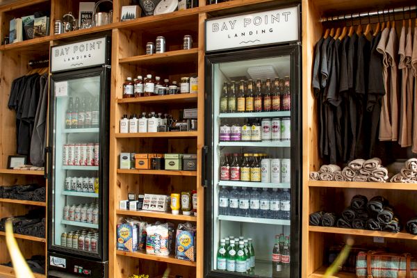 The image shows a shop interior with wooden shelves, refrigerators stocked with drinks, merchandise, and a plant in the foreground.