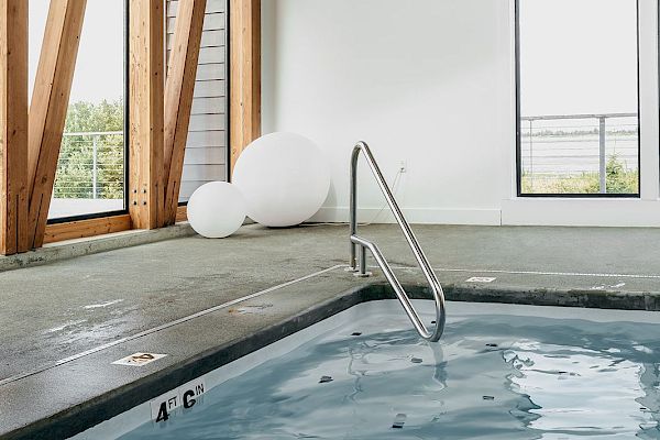 The image shows an indoor pool area with a handrail, windows, and modern wooden beams. Large white spherical decor is seen in the corner.