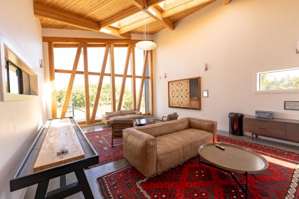 A modern living room with wooden beams, large windows, beige couches, a circular table, and red patterned rugs on the floor, completes the scene.