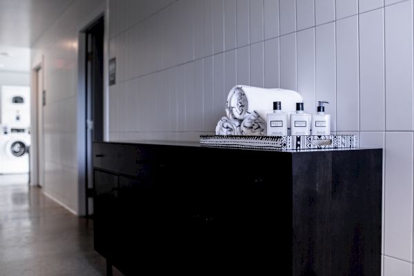 A hallway with a dark cabinet topped with rolled towels and toiletries. The background shows a partially visible laundry room with machines.